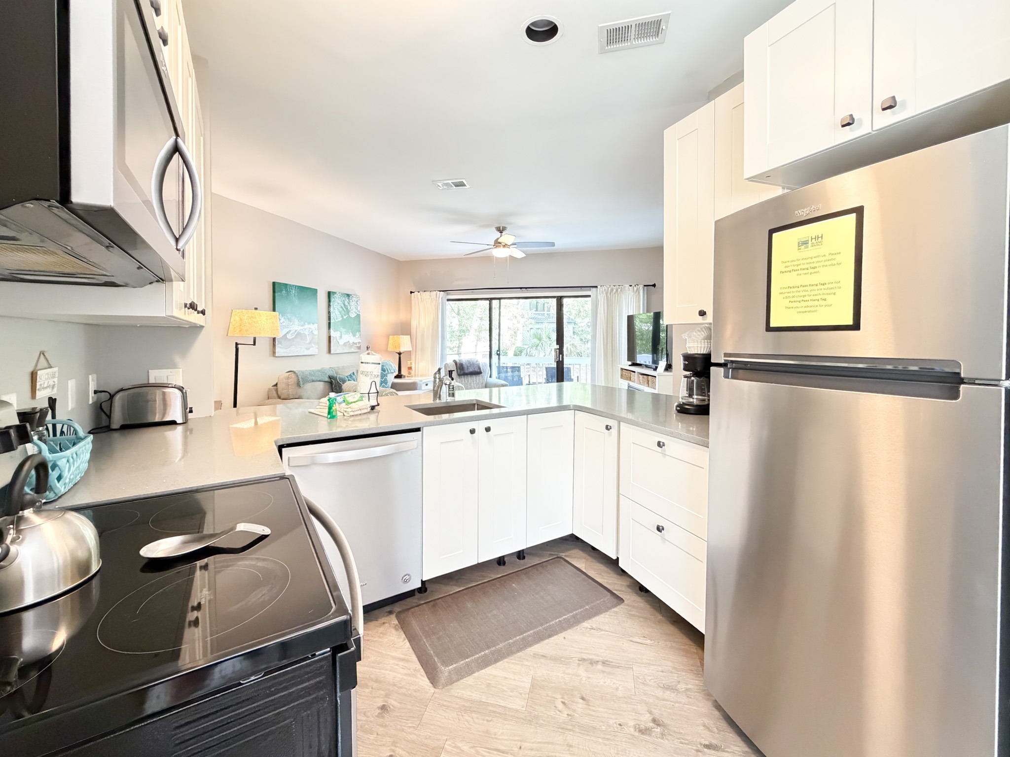Bright open kitchen with white cabinets, stainless-steel refrigerator, black electric stove, and island; living room visible through an opening in the background.