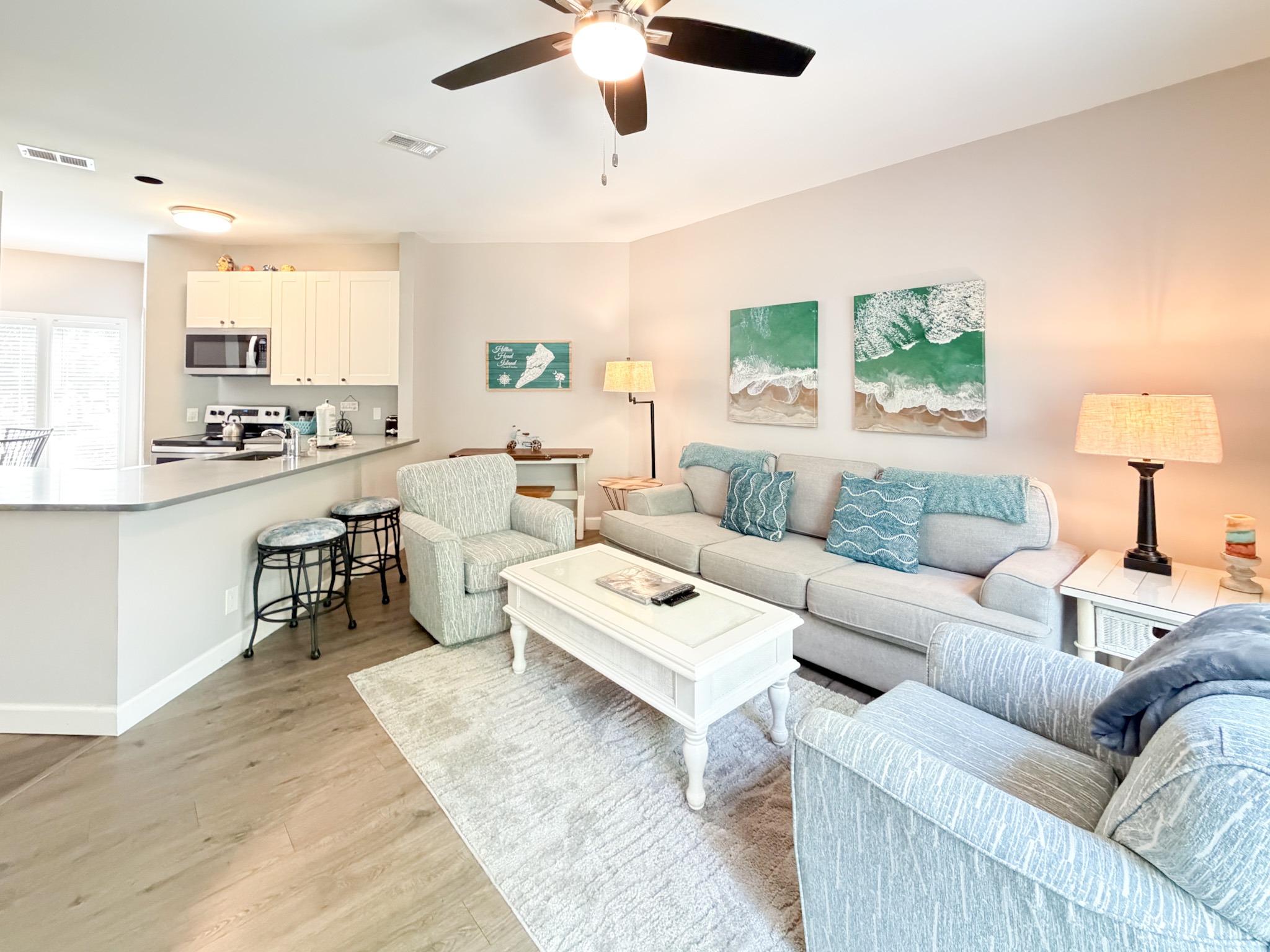 Open-concept living area with a gray sofa set, white coffee table, teal pillows, and coastal wall art; kitchen visible in the background.